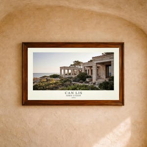May include: A rectangular wooden frame containing a horizontal photograph of the Can Lis stone house by Jørn Utzon, built in 1973. The image displays a coastal structure with open columns, stone walls, and surrounding greenery near the sea.