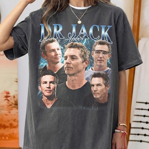 May include: Dark gray t-shirt featuring a collage of images of a man with the text "DR JACK Abbott" in blue. The shirt has a vintage, worn look.