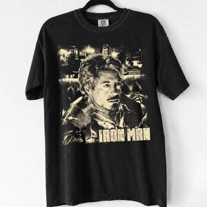 May include: A black short-sleeve t-shirt featuring a distressed, cream-colored graphic print of a man with a goatee. The text "IRON MAN" is printed in a bold, block font at the bottom of the graphic. The shirt is displayed on a black hanger.
