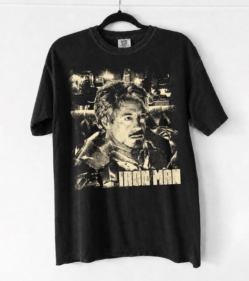 May include: A black short-sleeve t-shirt featuring a distressed, cream-colored graphic print of a man with a goatee. The text "IRON MAN" is printed in a bold, block font at the bottom of the graphic. The shirt is displayed on a black hanger.