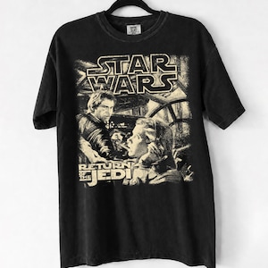 Pode incluir: Uma t-shirt preta de manga curta com uma impressão gráfica desgastada em tom creme de Han Solo e da Princesa Leia de Star Wars: O Regresso de Jedi. O design inclui o logótipo Star Wars no topo e o título do filme na parte inferior.