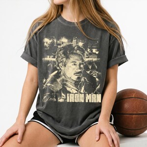 May include: A charcoal gray short-sleeve t-shirt featuring a distressed, light-colored graphic print of a man's face and the text IRON MAN centered on the chest. The shirt has a crew neckline and a relaxed fit with rolled sleeves.