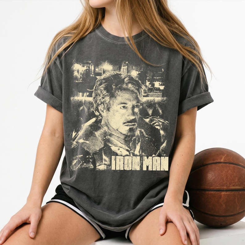 May include: A charcoal gray short-sleeve t-shirt featuring a distressed, light-colored graphic print of a man's face and the text IRON MAN centered on the chest. The shirt has a crew neckline and a relaxed fit with rolled sleeves.