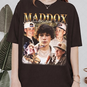May include: Dark brown t-shirt featuring a collage of images of a young man, with the name "MADDOX" in gold lettering above. The name "Batson" is written in cursive below. The shirt has a vintage, retro style.