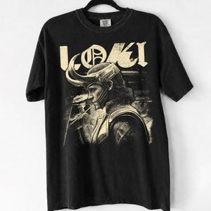 Op de afbeelding: Een zwart t-shirt met korte mouwen en een crèmekleurige grafische print van het personage Loki in profiel, met een gehoornde helm. De tekst LOKI is in een groot, verweerd lettertype boven de illustratie van het personage gedrukt.