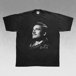 May include: Black t-shirt featuring a black and white portrait of a man looking upwards. The image includes the name "Reed Richards" in a handwritten font. The t-shirt is a crew neck style.