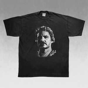 May include: Black t-shirt featuring a grayscale portrait of a man with a beard and signature. The image is centered on the shirt, showcasing the design. The shirt has a classic crew neck.