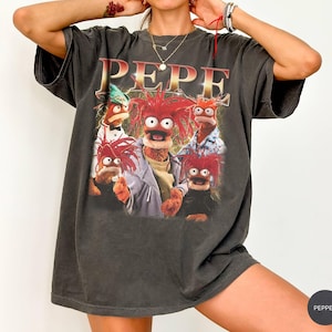 May include: Dark gray t-shirt featuring a graphic of the Muppets' Pepe the King Prawn and other characters. The word "PEPE" is printed in large, gold letters above the image. The shirt has a vintage, worn look.