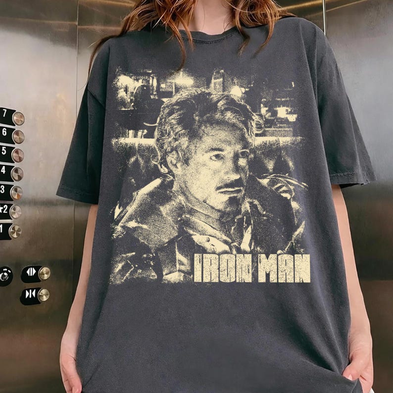 May include: A charcoal gray short-sleeve t-shirt featuring a distressed, light-colored graphic print of Tony Stark. The design includes a detailed portrait of the character and the text IRON MAN printed in a bold, block font at the bottom.