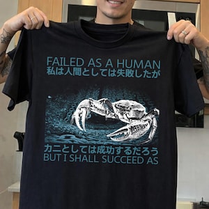 Könnte beinhalten: Schwarzes T-Shirt mit einem Krabben-Motiv und Text auf Englisch und Japanisch. Der Text lautet "FAILED AS A HUMAN" und "BUT I SHALL SUCCEED AS". Die Krabbengrafik ist in Graustufen gehalten.