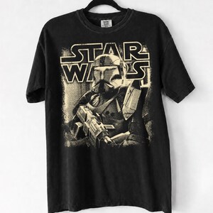 Peut inclure: Un t-shirt noir &agrave; manches courtes avec une impression graphique couleur cr&egrave;me repr&eacute;sentant un clone trooper Star Wars tenant un fusil blaster. Le texte STAR WARS est imprim&eacute; dans une police grasse et stylis&eacute;e au-dessus du personnage.
