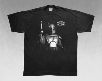 May include: Black t-shirt featuring a graphic of a character from Star Wars: Attack of the Clones. The character is wearing armor and holding a weapon. The text "STAR WARS ATTACK OF THE CLONES EPISODE II" is printed on the shirt.