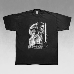 May include: Black t-shirt featuring a grayscale graphic from Star Wars: Attack of the Clones. The design includes characters and the movie title. The shirt has a crew neck and short sleeves.