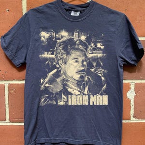 May include: A navy blue short-sleeve t-shirt featuring a light-colored graphic print of a man's face and the text "IRON MAN" in a bold, block font. The graphic has a distressed, vintage-style appearance and is centered on the front of the shirt.