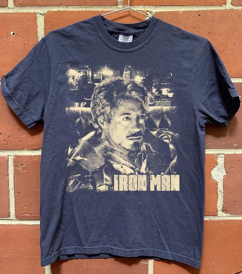 May include: A navy blue short-sleeve t-shirt featuring a light-colored graphic print of a man's face and the text "IRON MAN" in a bold, block font. The graphic has a distressed, vintage-style appearance and is centered on the front of the shirt.