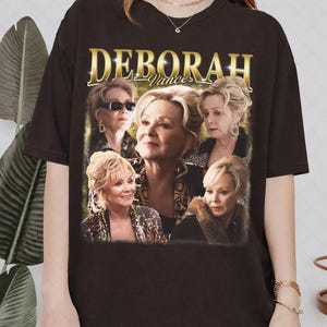 May include: Dark brown t-shirt featuring a gold-colored text that reads "DEBORAH Vance" above a collage of images of a woman with blonde hair. The images show the woman in various poses.