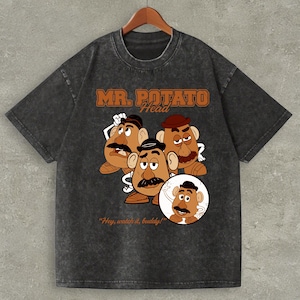 May include: Dark gray t-shirt featuring a graphic of Mr. Potato Head characters. The design includes the words "MR. POTATO Head" in orange lettering and the phrase "Hey, watch it, buddy!" in white. The shirt has a vintage, distressed look.