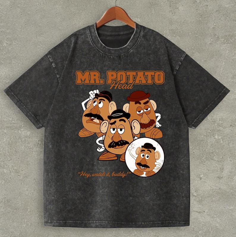 May include: Dark gray t-shirt featuring a graphic of Mr. Potato Head characters. The design includes the words "MR. POTATO Head" in orange lettering and the phrase "Hey, watch it, buddy!" in white. The shirt has a vintage, distressed look.