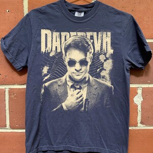 Peut inclure: Un t-shirt bleu marine à manches courtes avec une impression graphique couleur crème représentant une personne en costume portant des lunettes de soleil rondes. Le texte DAREDEVIL est imprimé au-dessus avec un fond de gratte-ciel.