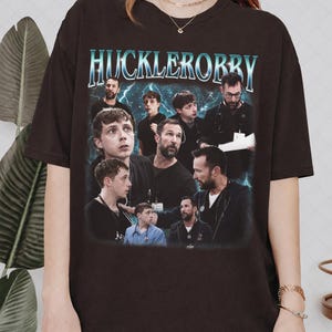 May include: A dark brown t-shirt with the text "HUCKLEORRBY" in teal, distressed lettering. The shirt displays a collage of images of people, set against a backdrop of a stormy sky, creating a vintage aesthetic.