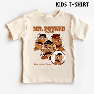 May include: A cream-colored kids t-shirt featuring a graphic of Mr. Potato Head characters. The design includes the text "MR. POTATO Head" in brown lettering, along with the phrase "Hey, watch it, buddy!" The shirt is short-sleeved.