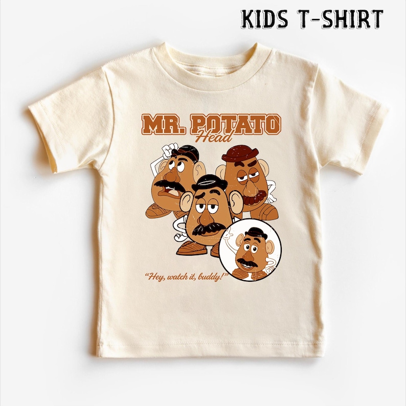 May include: A cream-colored kids t-shirt featuring a graphic of Mr. Potato Head characters. The design includes the text "MR. POTATO Head" in brown lettering, along with the phrase "Hey, watch it, buddy!" The shirt is short-sleeved.