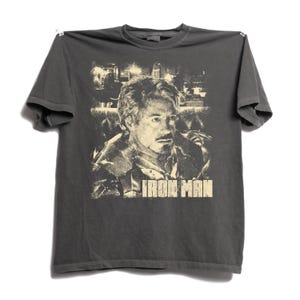 May include: A charcoal gray short-sleeve t-shirt featuring a cream-colored graphic print of a person with a goatee. The graphic includes a detailed portrait and the text IRON MAN in a bold, blocky font centered below the portrait on the chest.