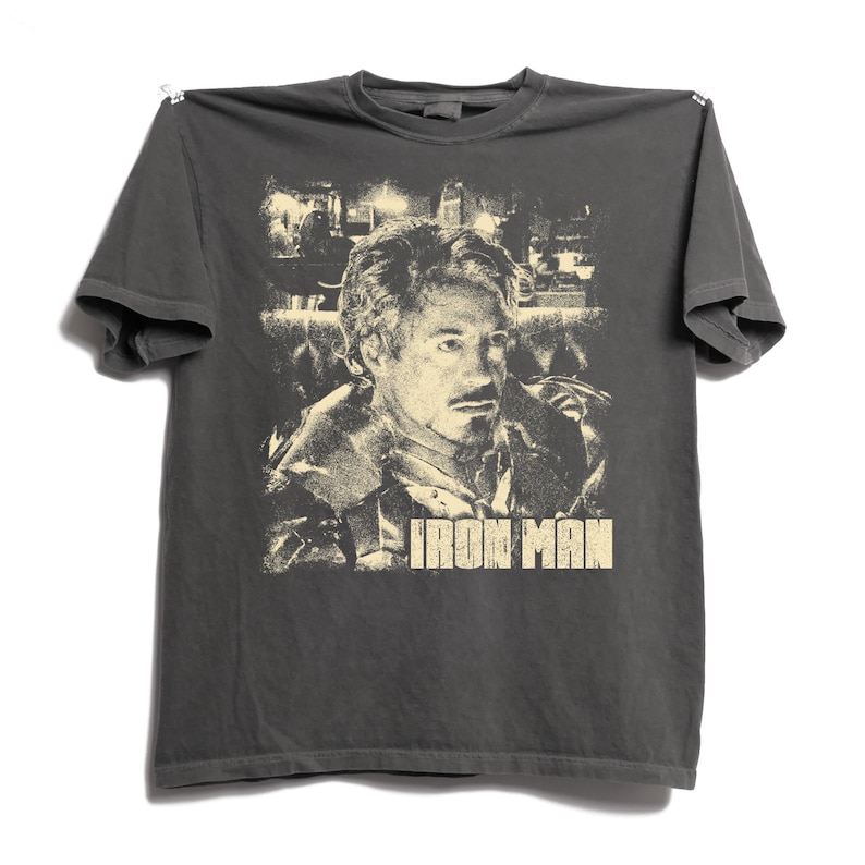 May include: A charcoal gray short-sleeve t-shirt featuring a cream-colored graphic print of a person with a goatee. The graphic includes a detailed portrait and the text IRON MAN in a bold, blocky font centered below the portrait on the chest.