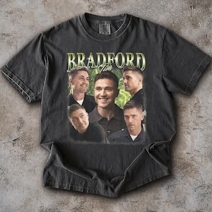 May include: Dark grey t-shirt with a vintage-style graphic. The graphic features the name "Bradford" and the name "Tim" in green font, along with multiple images of a man. The shirt is displayed flat on a textured surface.