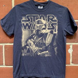 Peut inclure: Un t-shirt bleu marine &agrave; manches courtes avec une impression graphique couleur cr&egrave;me repr&eacute;sentant un clone trooper Star Wars tenant un fusil blaster. Le texte STAR WARS est imprim&eacute; dans une police grasse et d&eacute;tour&eacute;e au-dessus du personnage.