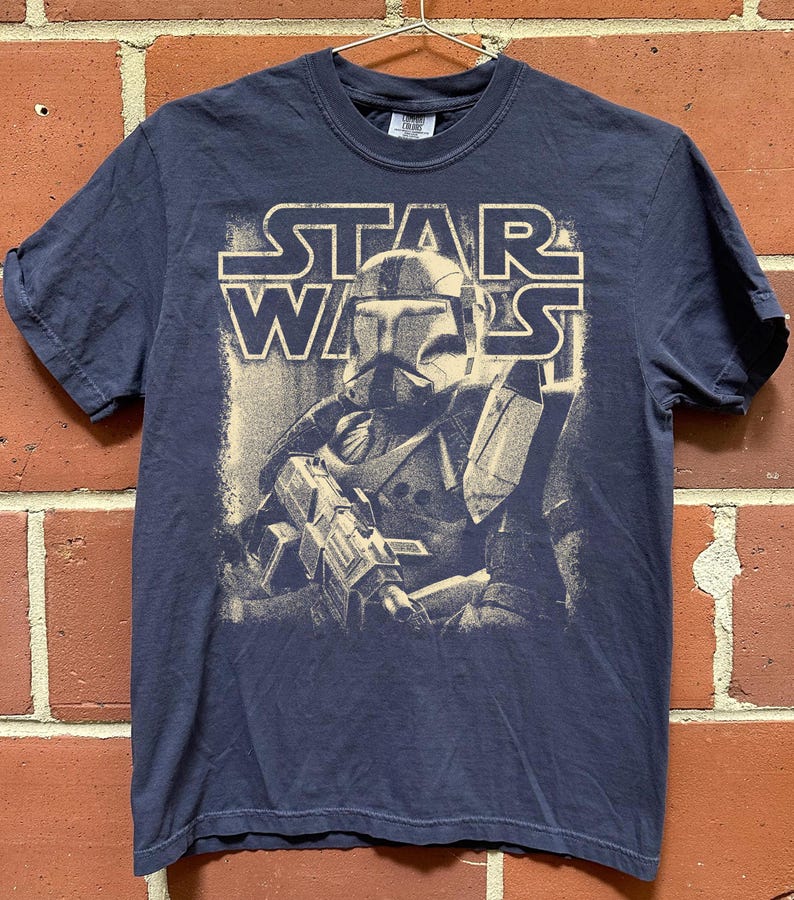 Peut inclure: Un t-shirt bleu marine &agrave; manches courtes avec une impression graphique couleur cr&egrave;me repr&eacute;sentant un clone trooper Star Wars tenant un fusil blaster. Le texte STAR WARS est imprim&eacute; dans une police grasse et d&eacute;tour&eacute;e au-dessus du personnage.