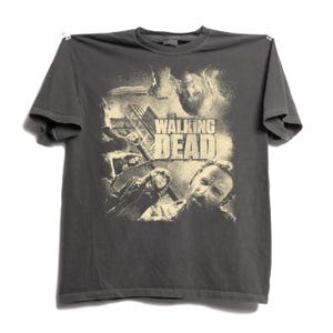 May include: A charcoal gray short-sleeve t-shirt featuring a cream-colored graphic print of characters from The Walking Dead. The design includes the show title in bold lettering, a building structure, and several figures in a distressed style.