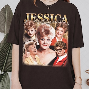Puede incluir: Camiseta marrón oscuro con un collage de imágenes de Jessica Fletcher, con el nombre "JESSICA" en letras doradas sobre las imágenes. La palabra "Fletcher" está debajo del nombre. Las imágenes muestran a Jessica Fletcher en varias poses.