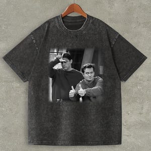 May include: Dark grey t-shirt with a black and white image of two men. One man is making a loser sign, the other gives a thumbs up. The shirt is displayed on a wooden hanger.