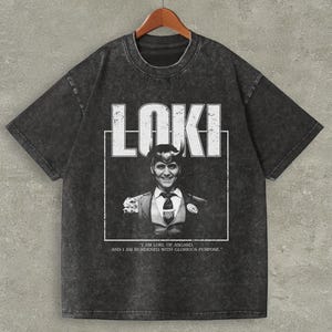 以下が含まれることがあります： 「LOKI」の文字が白でプリントされた濃いグレーのTシャツ。テキストの下には、スーツを着た人物の白黒画像があります。「I AM LOKI, OF ASGARD, AND I AM BURDENED WITH GLORIOUS PURPOSE.」という引用句が画像の下に印刷されています。