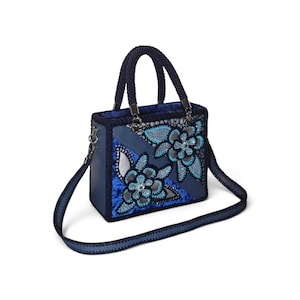May include: A structured navy blue handbag featuring a decorative floral appliqu&eacute; in shades of bright blue, silver, and black. The bag includes a braided top handle, a detachable shoulder strap, and silver-toned hardware accents on the sides.