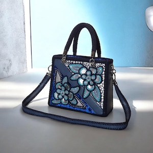 May include: A structured handbag in shades of blue, featuring a dark blue braided handle and a matching detachable shoulder strap. The front panel displays intricate silver and turquoise floral embroidery with sparkling accents and lace detailing.