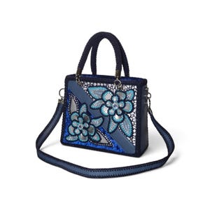 May include: A structured handbag in shades of blue and silver featuring a dark blue braided handle and a matching detachable shoulder strap. The front panel displays a floral pattern with silver accents, blue sequins, and intricate lace detailing.