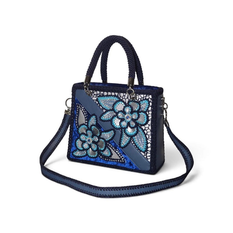 May include: A structured handbag in shades of blue and silver featuring a dark blue braided handle and a matching detachable shoulder strap. The front panel displays a floral pattern with silver accents, blue sequins, and intricate lace detailing.