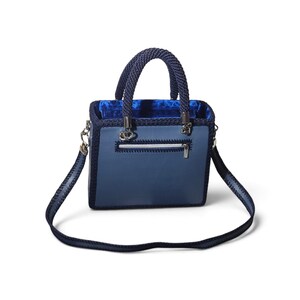 May include: A structured square handbag in shades of blue, featuring a smooth leather-like front panel with a horizontal zippered pocket, dark blue crocheted trim, two rounded braided handles, and a matching detachable shoulder strap.