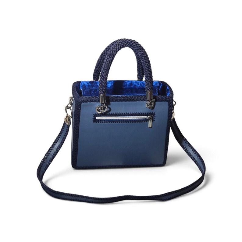 May include: A structured square handbag in shades of blue, featuring a smooth leather-like front panel with a horizontal zippered pocket, dark blue crocheted trim, two rounded braided handles, and a matching detachable shoulder strap.