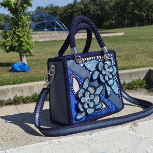 May include: A structured handbag featuring a dark blue crocheted border and handles, with a grey base and decorative floral appliqu&eacute;s in shades of blue, light blue, and silver. The bag includes a matching detachable shoulder strap with silver hardware.