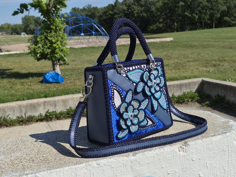May include: A structured handbag featuring a dark blue crocheted border and handles, with a grey base and decorative floral appliqu&eacute;s in shades of blue, light blue, and silver. The bag includes a matching detachable shoulder strap with silver hardware.