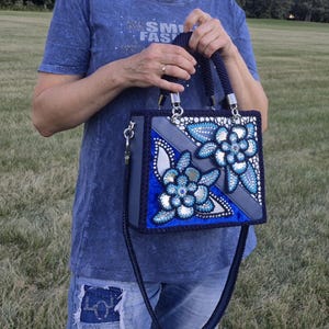 May include: A structured rectangular handbag featuring a deep blue and grey color palette with decorative floral appliques. The bag includes two braided dark blue handles, silver hardware, and a long matching shoulder strap with crystal accents.