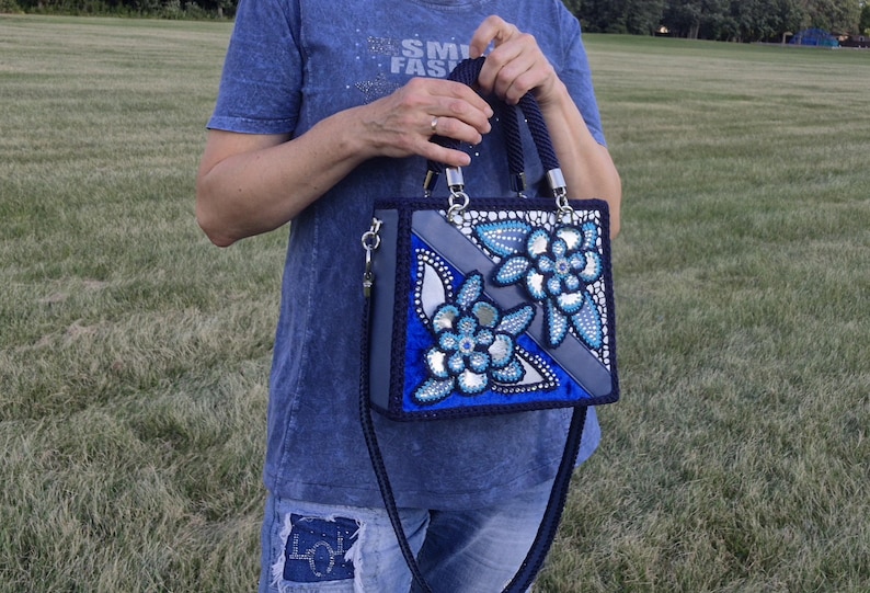 May include: A structured rectangular handbag featuring a deep blue and grey color palette with decorative floral appliques. The bag includes two braided dark blue handles, silver hardware, and a long matching shoulder strap with crystal accents.