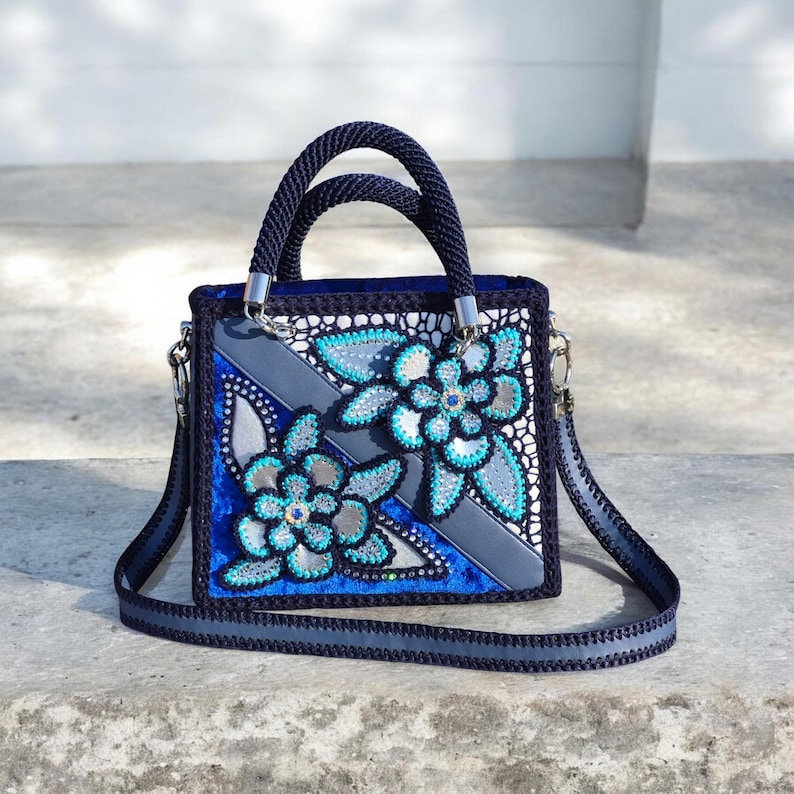 May include: A square-shaped handbag featuring a dark blue crocheted exterior with two top handles and a detachable shoulder strap. The front panel displays a decorative pattern of two large floral motifs in turquoise, silver, and dark blue beads.