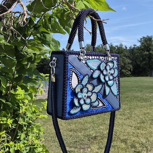 May include: A structured navy blue handbag featuring a crochet border and two top handles. The front panel displays a geometric design with blue and light blue floral appliqu&eacute;s embellished with sparkling rhinestones and intricate beadwork.
