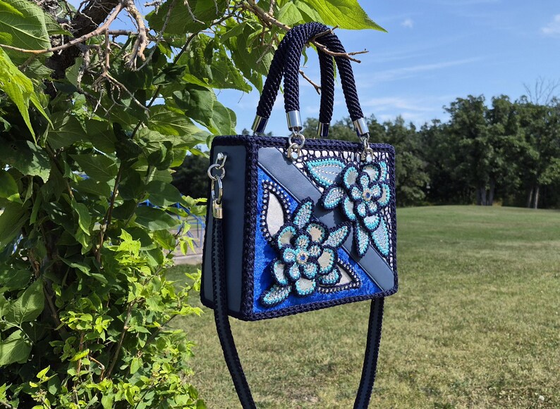 May include: A structured navy blue handbag featuring a crochet border and two top handles. The front panel displays a geometric design with blue and light blue floral appliqu&eacute;s embellished with sparkling rhinestones and intricate beadwork.