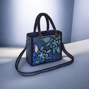 May include: A structured handbag in shades of blue, featuring a dark blue braided handle and a detachable shoulder strap. The exterior displays a combination of smooth blue panels, textured blue velvet, and intricate floral lace appliqu&eacute;s with silver and turquoise accents.