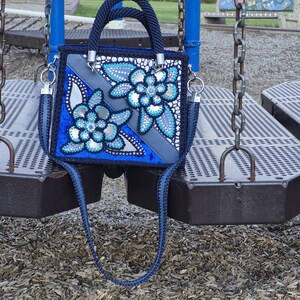 May include: A square-shaped navy blue handbag featuring a decorative floral pattern with silver and light blue accents. The bag includes a braided navy blue handle, a matching shoulder strap, and silver-toned hardware attachments on the sides.